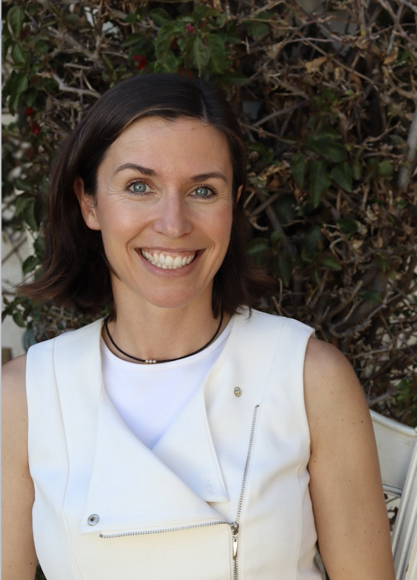 A smiling woman with short dark hair and blue eyes, wearing a white sleeveless top, outdoors with green and brown foliage in the background.