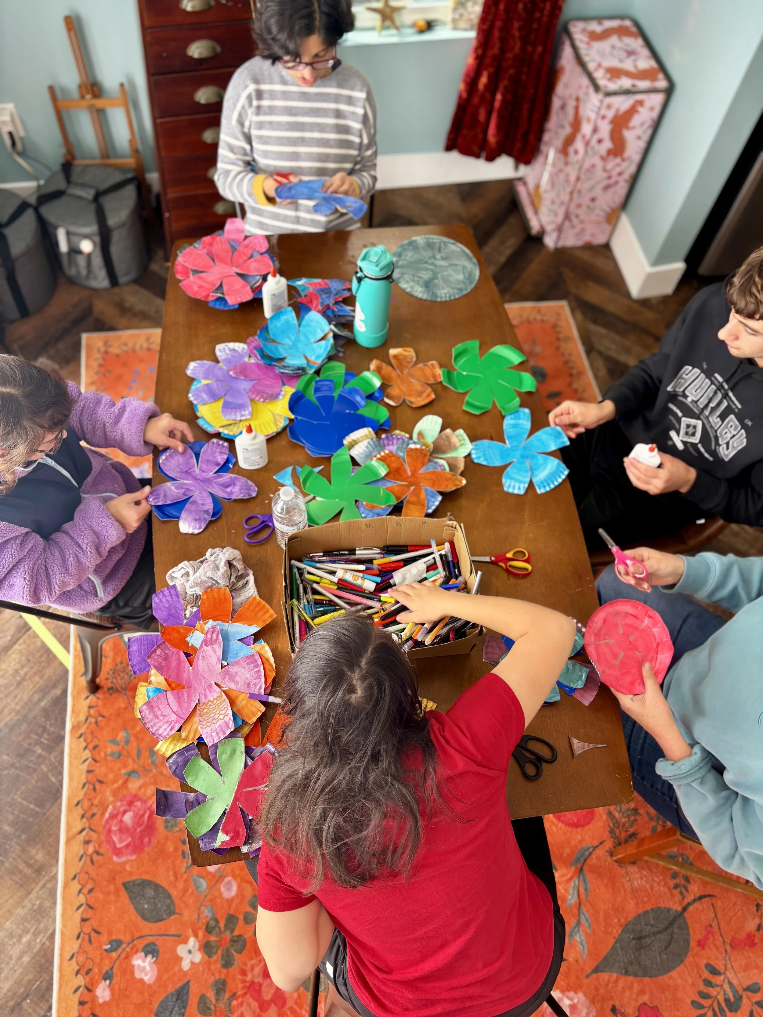 Children engaging in an arts and crafts activity at a wooden table, creating colorful paper flowers with scissors, glue, and markers.