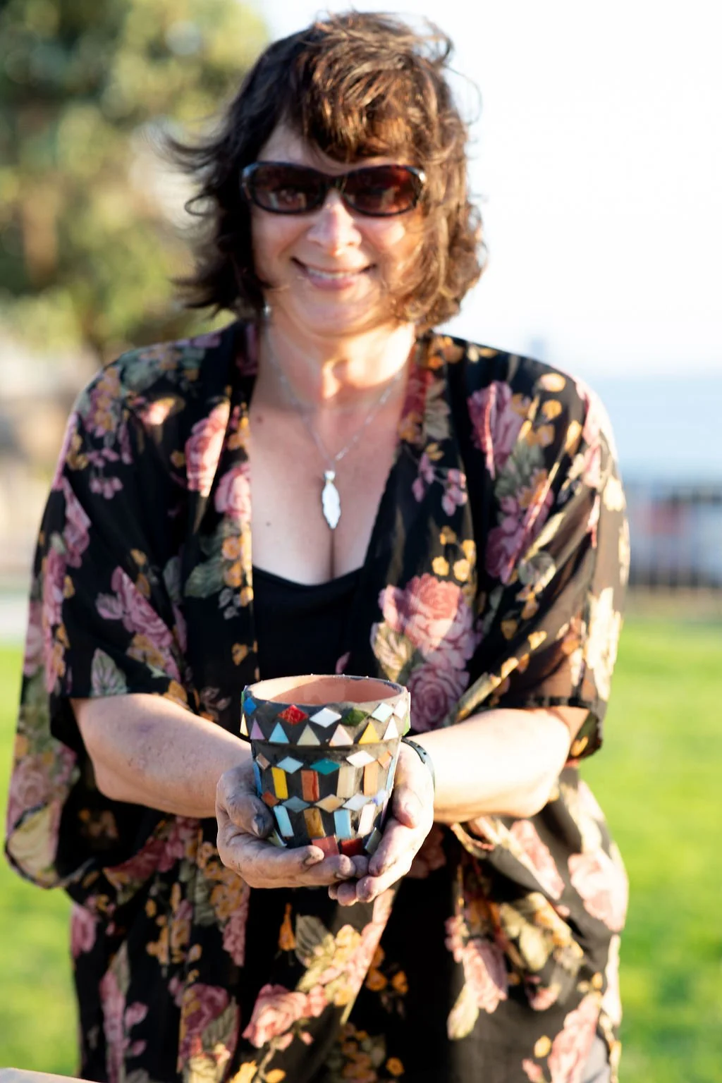 A smiling woman wearing sunglasses and a floral kimono-style jacket holds a colorful mosaic cup outdoors, with blurred greenery and water in the background.