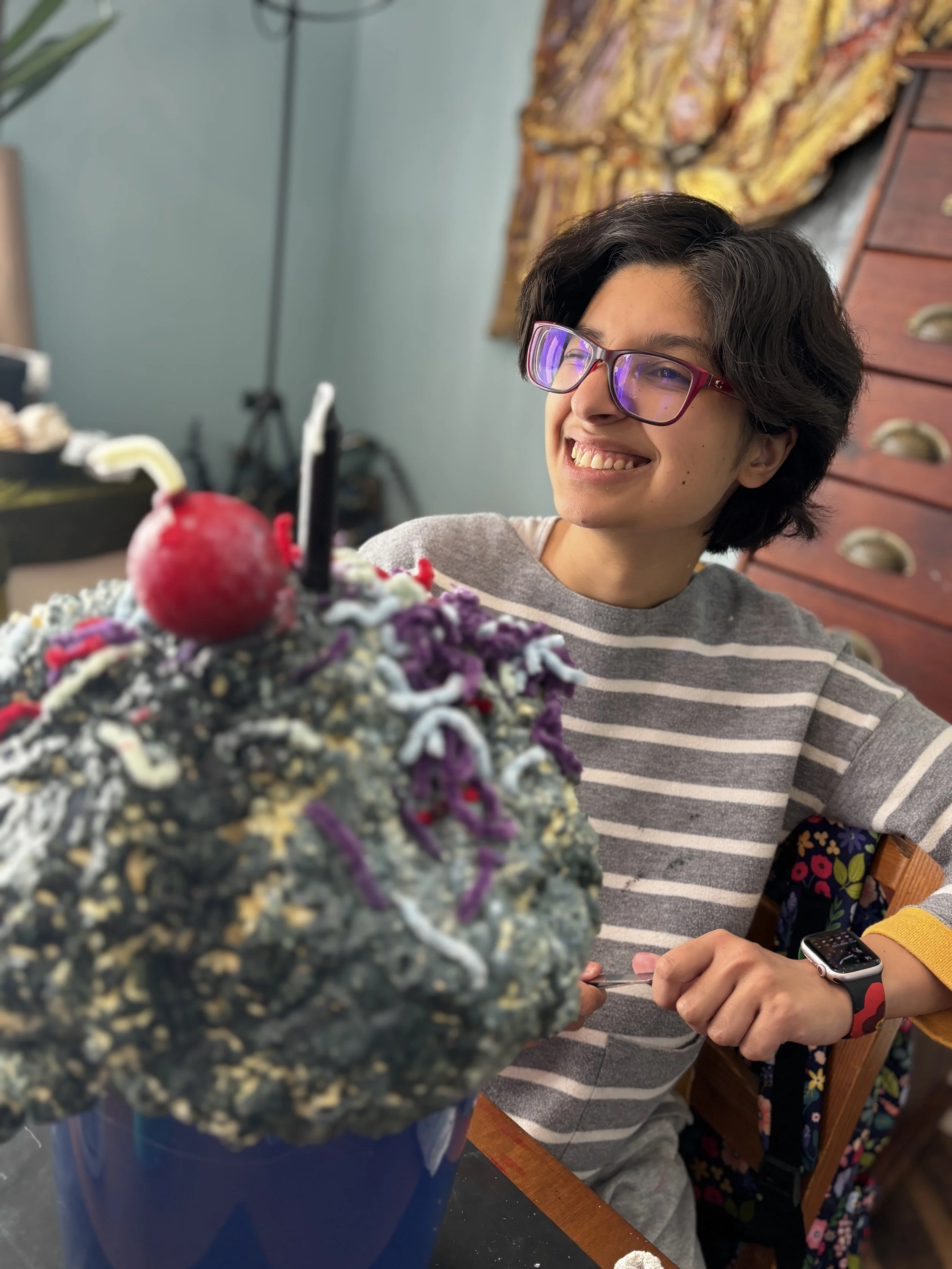 Person with short dark hair, glasses, and a striped gray and white shirt smiling at a birthday cake with a cherry and candles.