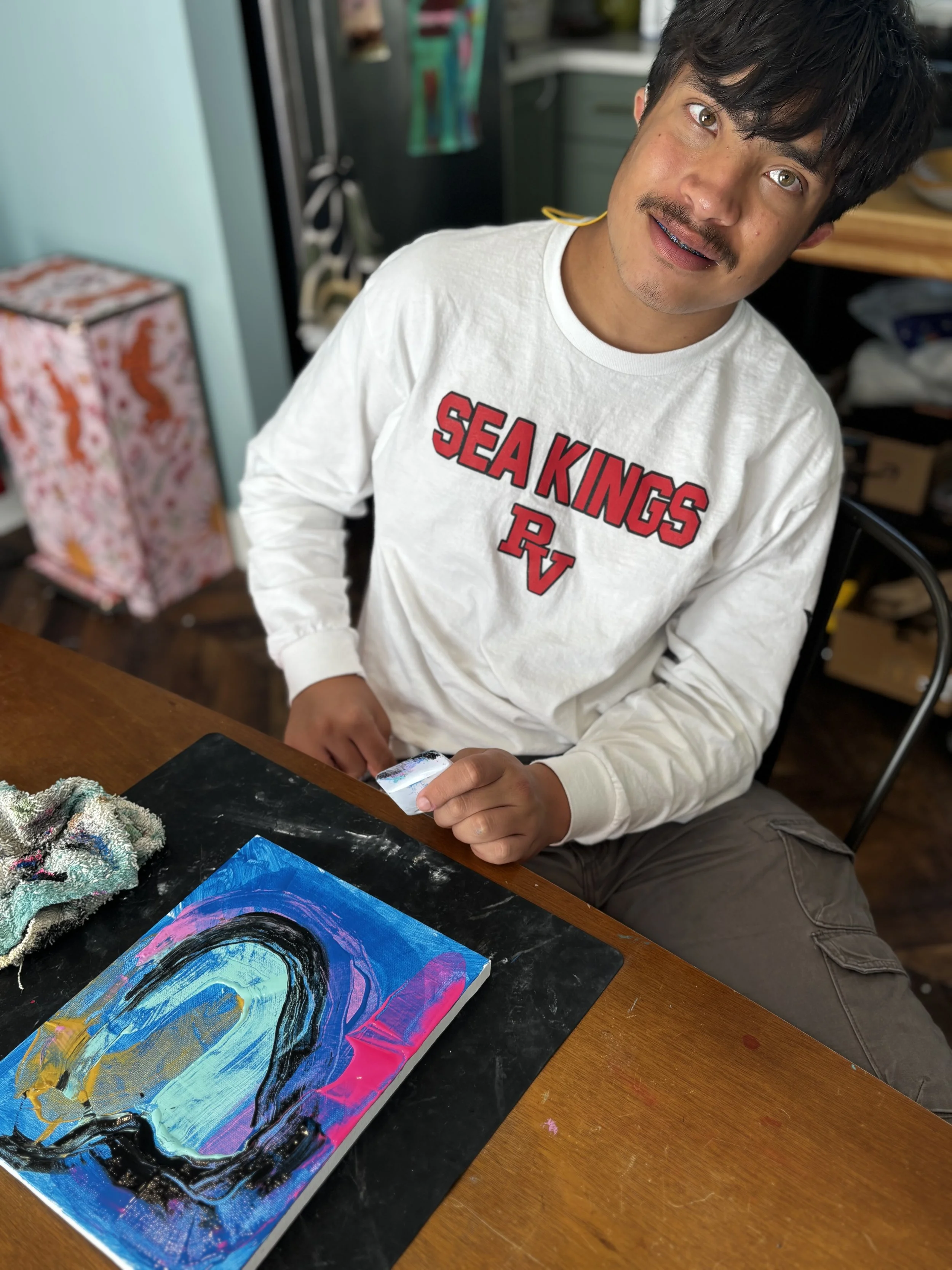 A young man with dark hair and a mustache, wearing a white sweatshirt with red text "SEA KINGS RV," sitting at a wooden table with colorful paint on canvas and a rag nearby, in a room with shelves and a floral gift bag in the background.