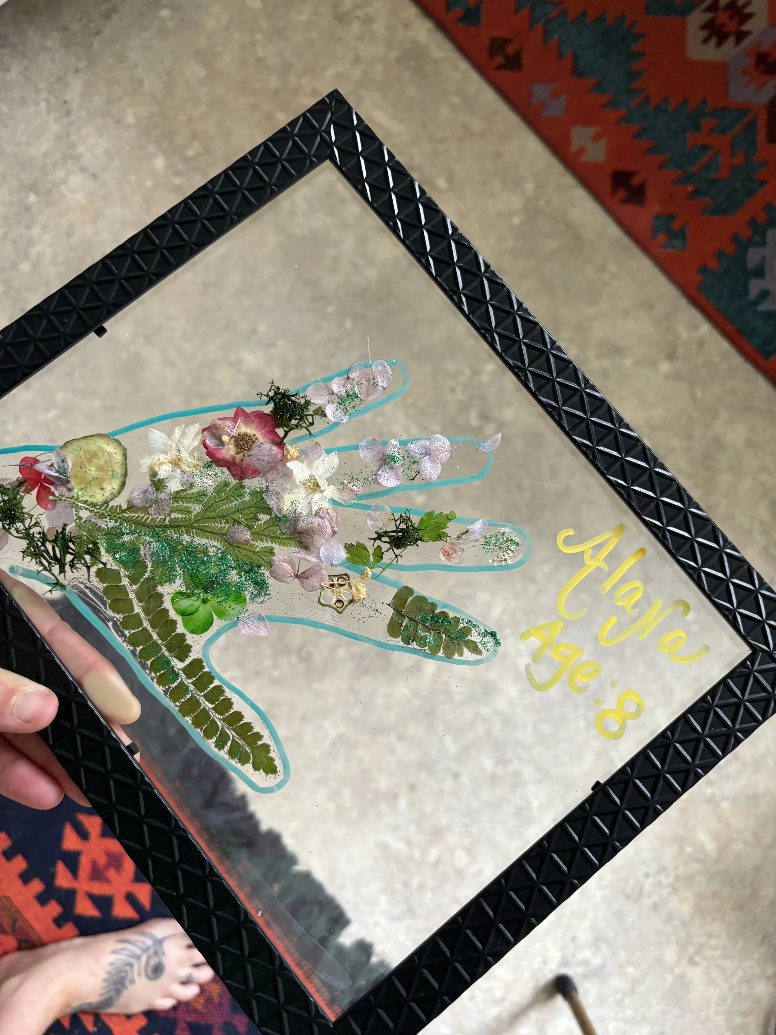 Framed artwork of a hand made from various pressed flowers, leaves, and natural materials, with the name 'Alaia' and age '8' written in yellow on the glass.