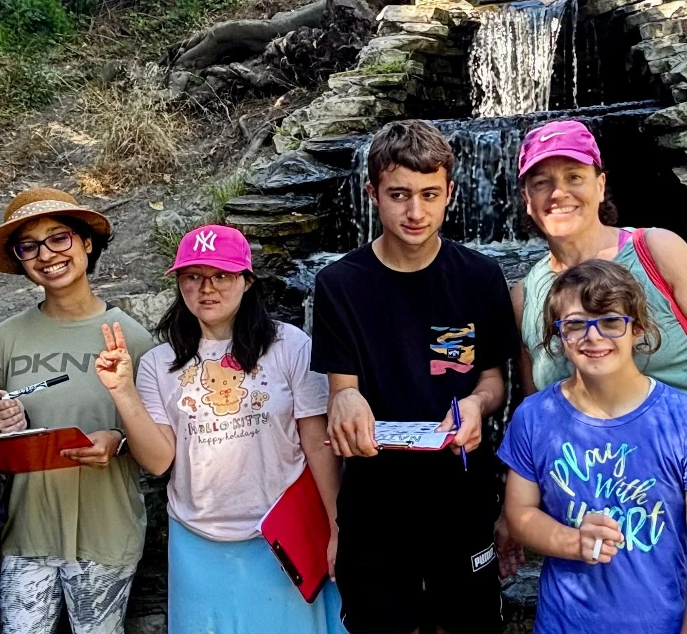 Group of six people standing in front of a small waterfall, smiling and posing outdoors. Four women and two young girls, with casual clothing and hats.