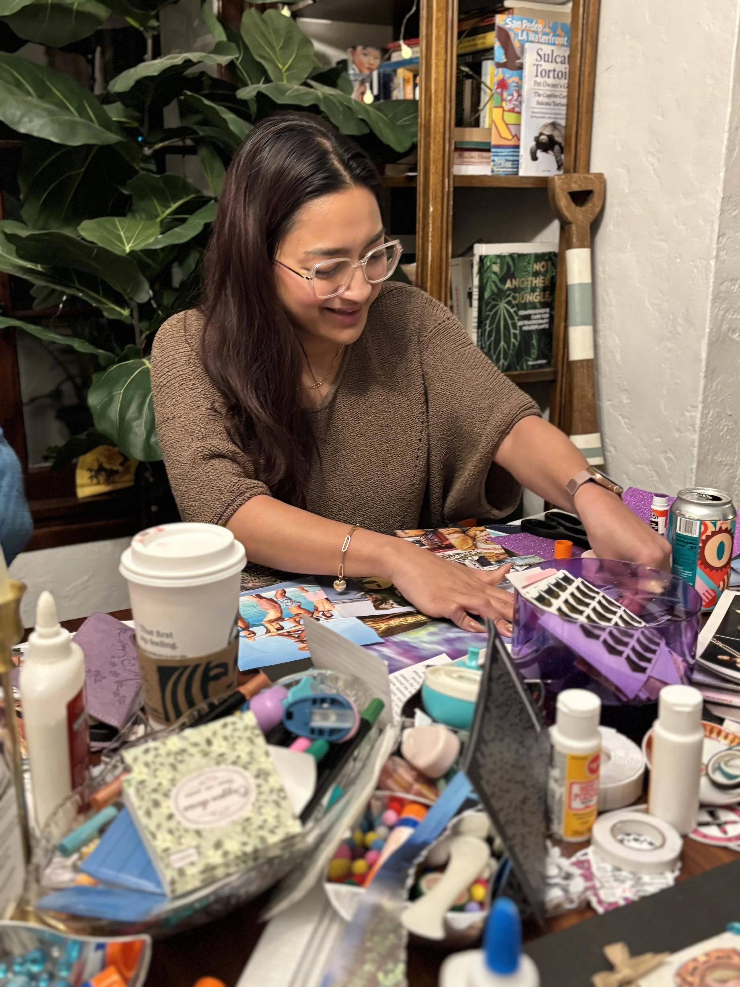 A woman with long dark hair and glasses is sitting at a cluttered table filled with craft supplies, including glue, scissors, markers, and paper. She is smiling and working on a craft project in a cozy room with bookshelves, plants, and decorative it