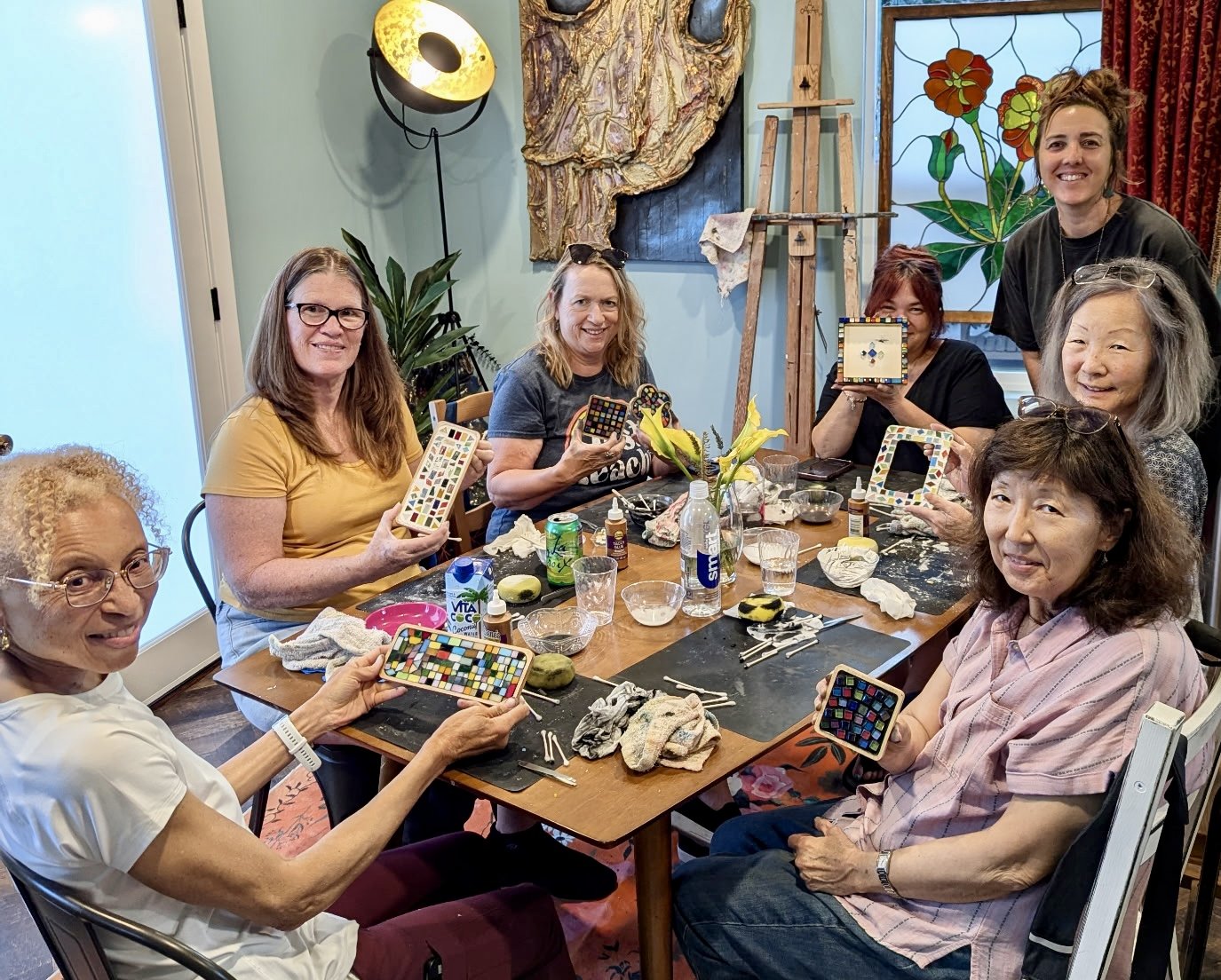 Group of women sitting around a table holding mosaic-patterned objects, engaging in an arts and crafts activity in a cozy, decorated room.