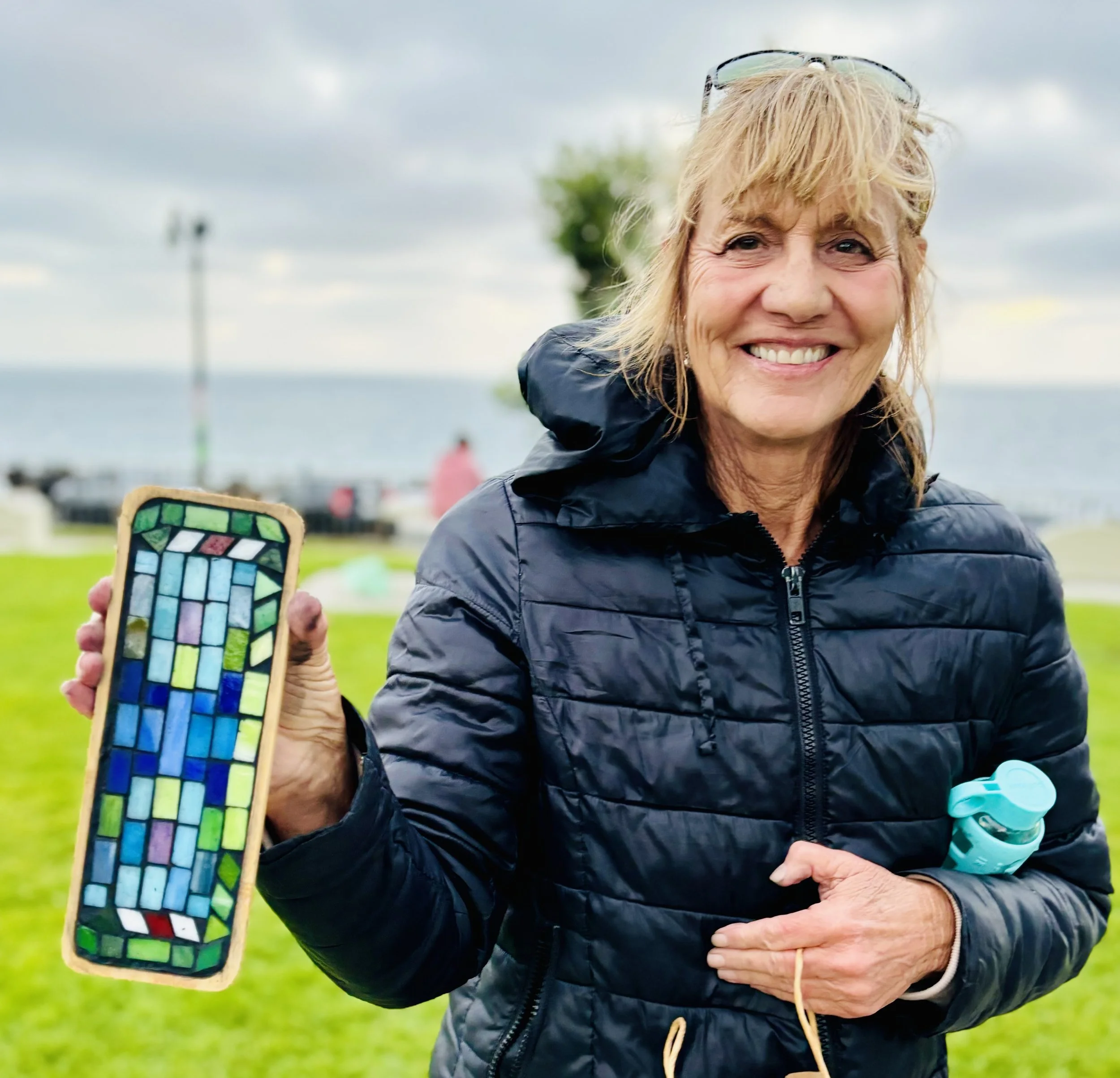 A smiling woman in a dark blue jacket holding a colorful stained glass mosaic in a wooden frame outdoors near the water.
