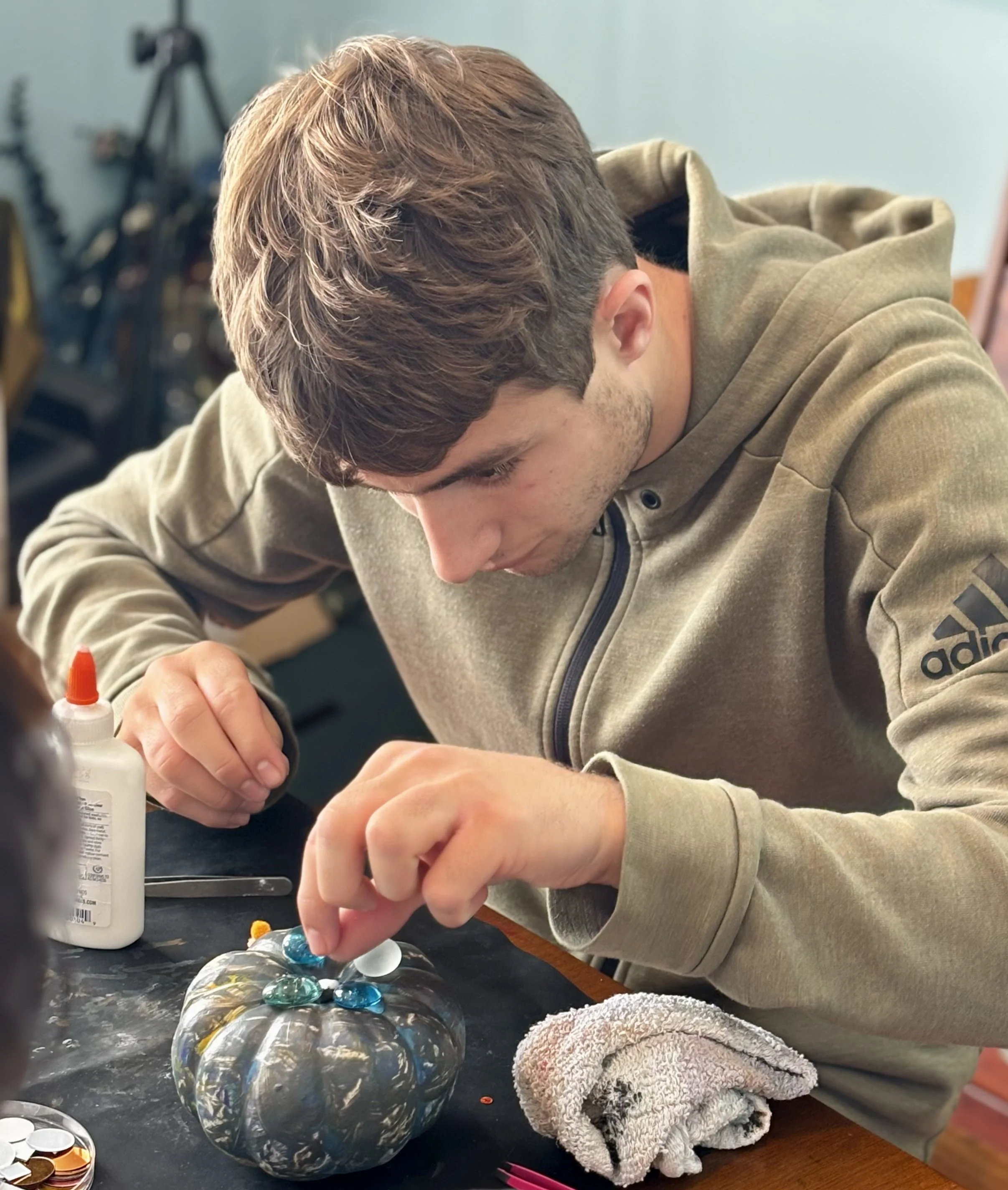 A young man with short brown hair, wearing a beige Adidas hoodie, is decorating a small pumpkin with blue and white glass marbles and glue. He is focused on his craft project at a table with supplies, including glue, a towel, and coins, in a room wit
