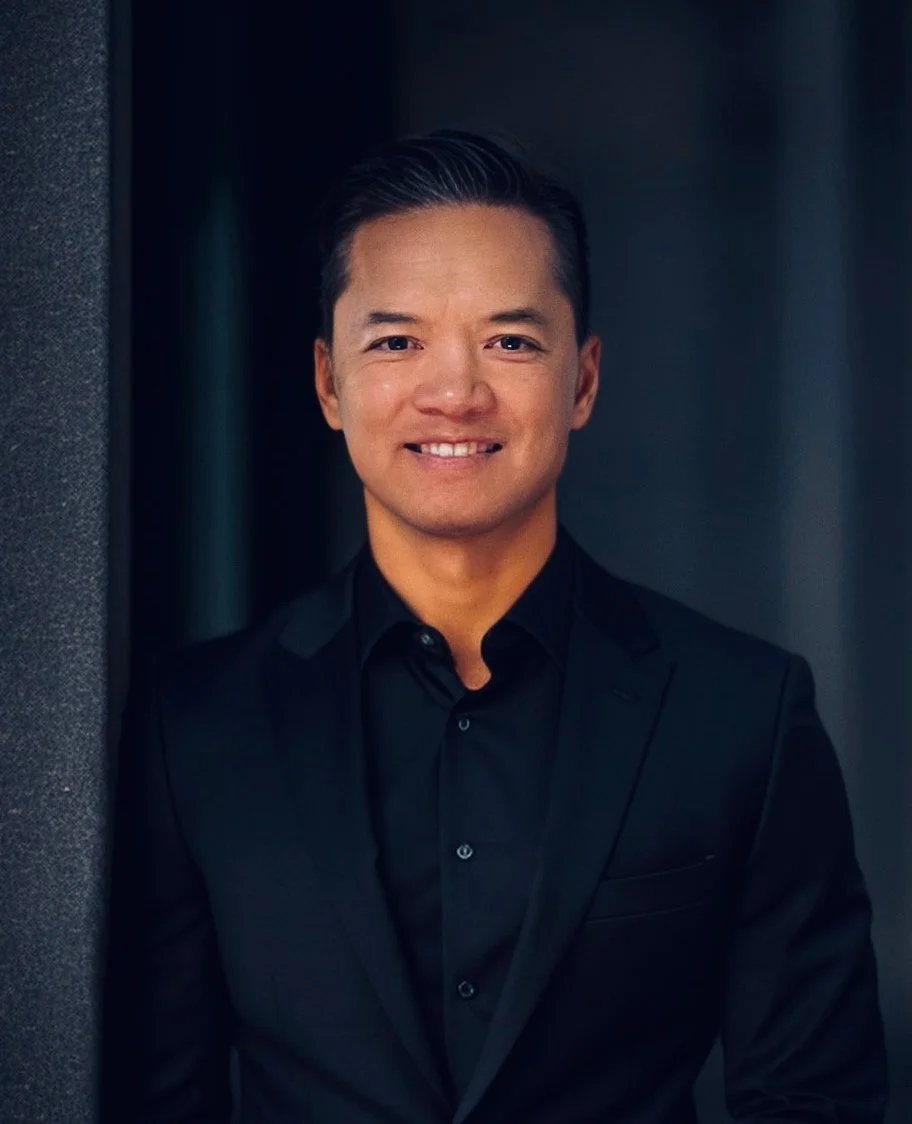 A man smiling, wearing a black suit and black shirt, standing in front of a dark background.