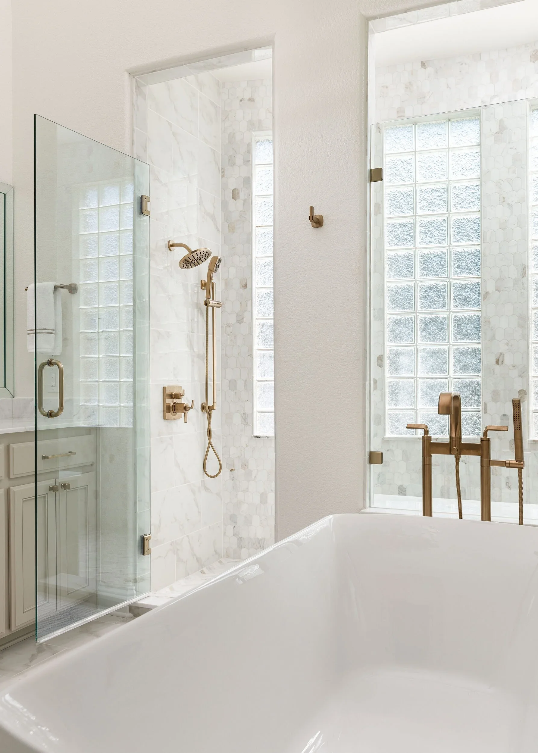 Glass shower enclosure beside a freestanding tub with a brass floor-mount faucet, built by Agapé Home Services.