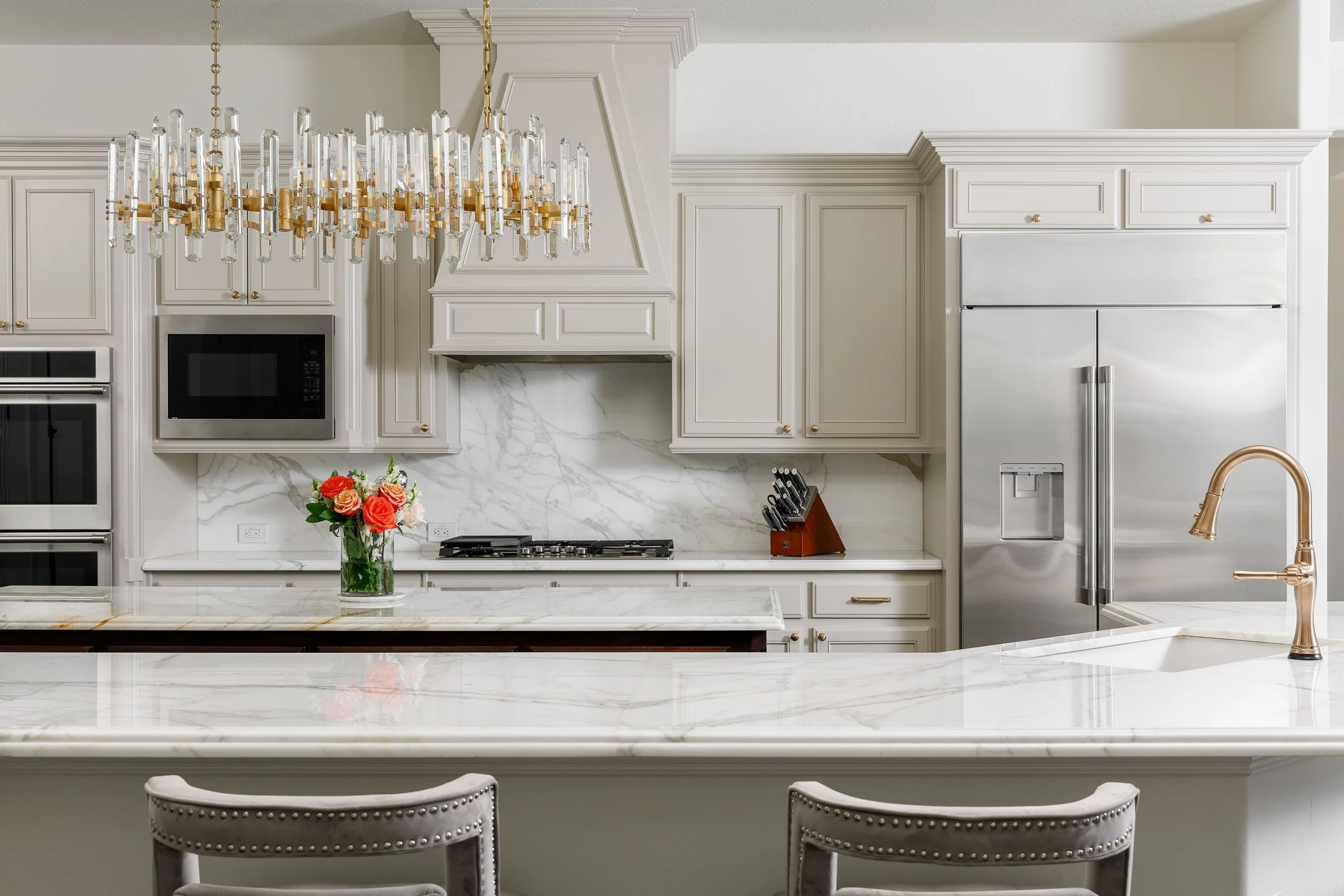 A modern kitchen with white cabinets, marble countertops, a stainless steel refrigerator, and a gold and glass chandelier. There is a vase of pink and orange roses on the island and a knife block on the counter.