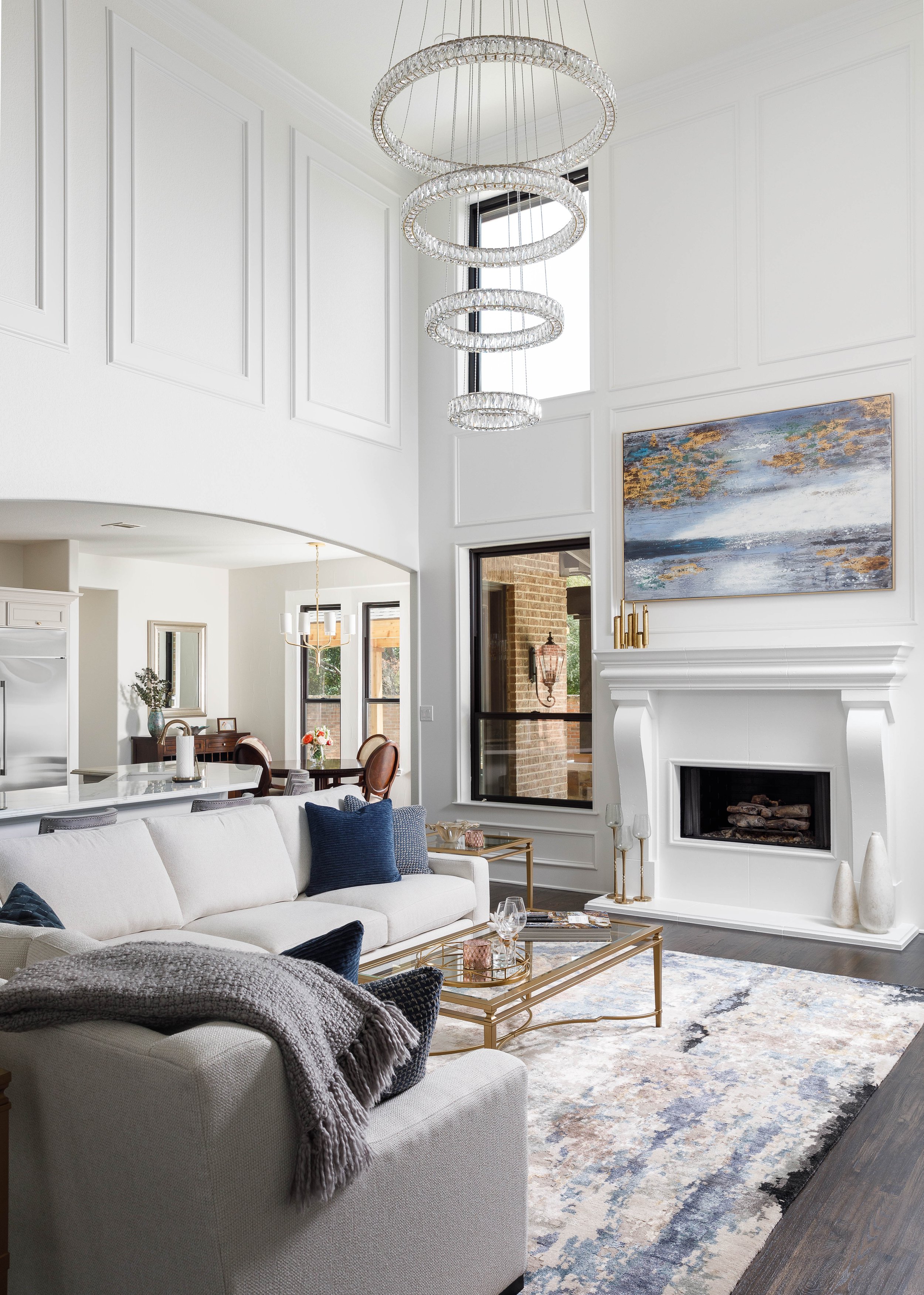 Modern living room with white walls, high ceiling, and a large window. Features a white sofa with navy and black pillows, a glass coffee table, and a fireplace with artwork above. A contemporary chandelier hangs from the ceiling.