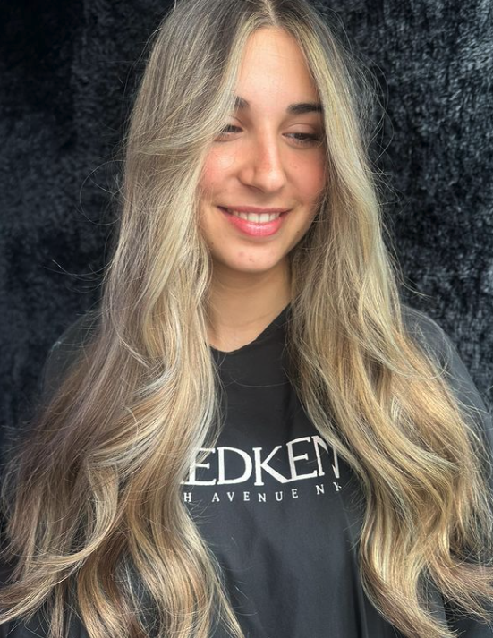 A young woman with long, wavy blonde hair smiling and looking down, wearing a black hoodie with white text, standing in front of a dark textured backdrop.