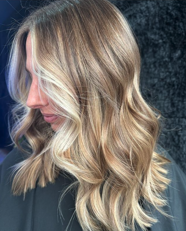 Close-up of a woman with shoulder-length wavy blonde hair styled with soft curls and highlights, looking down and wearing a dark top.
