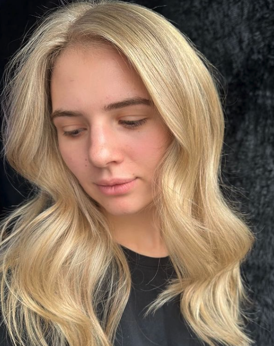 Close-up of a young woman with long, wavy blonde hair and fair skin, looking downward, against a black textured background.