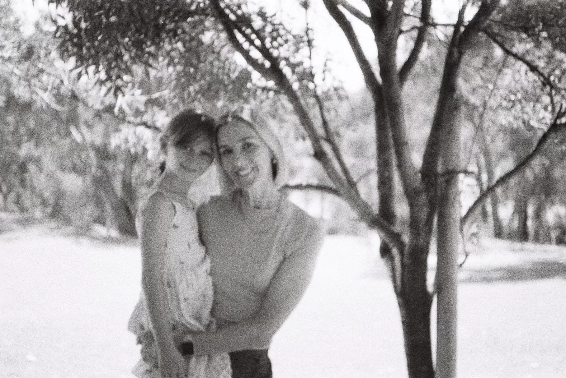 A woman and a young girl smiling and hugging outdoors near a tree.
