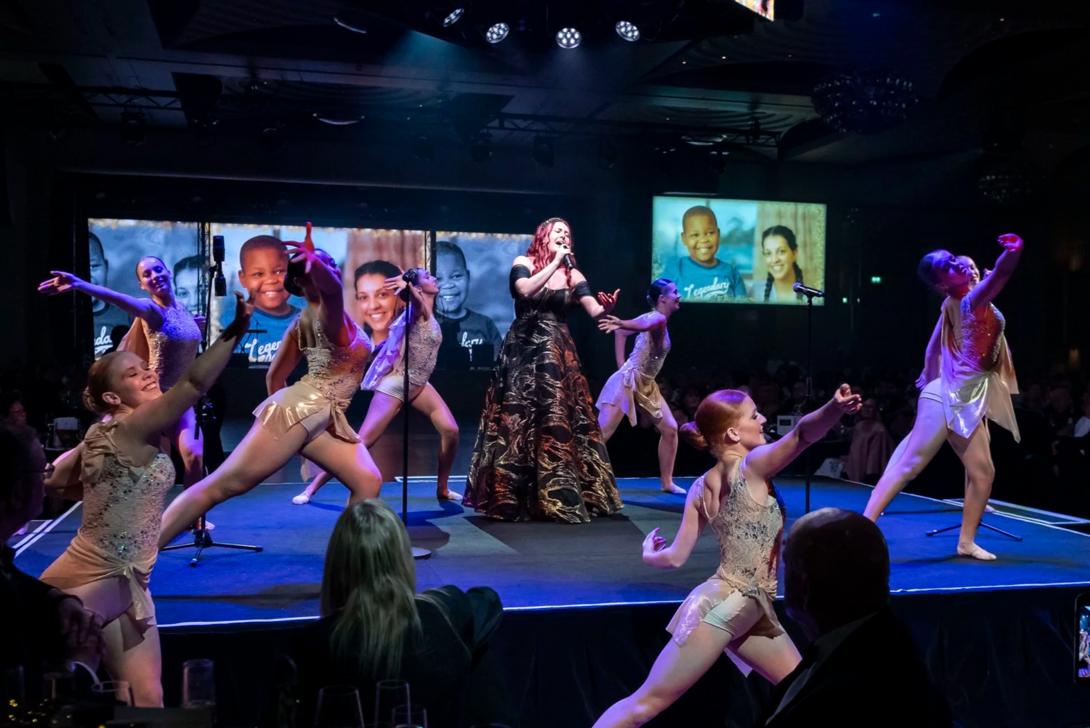 A stage performance featuring a woman singing into a microphone, surrounded by young dancers in gold costumes performing dance moves. Two large screens in the background display photographs of children.