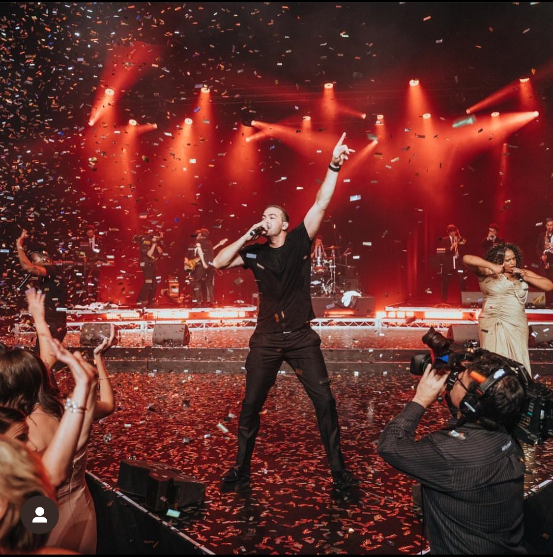 Performer singing on stage with red lighting, confetti falling, audience capturing the moment, and a live band playing in the background.