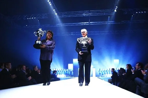 Two women standing on a runway stage holding large trophies, with an audience seated on either side, under bright stage lights.