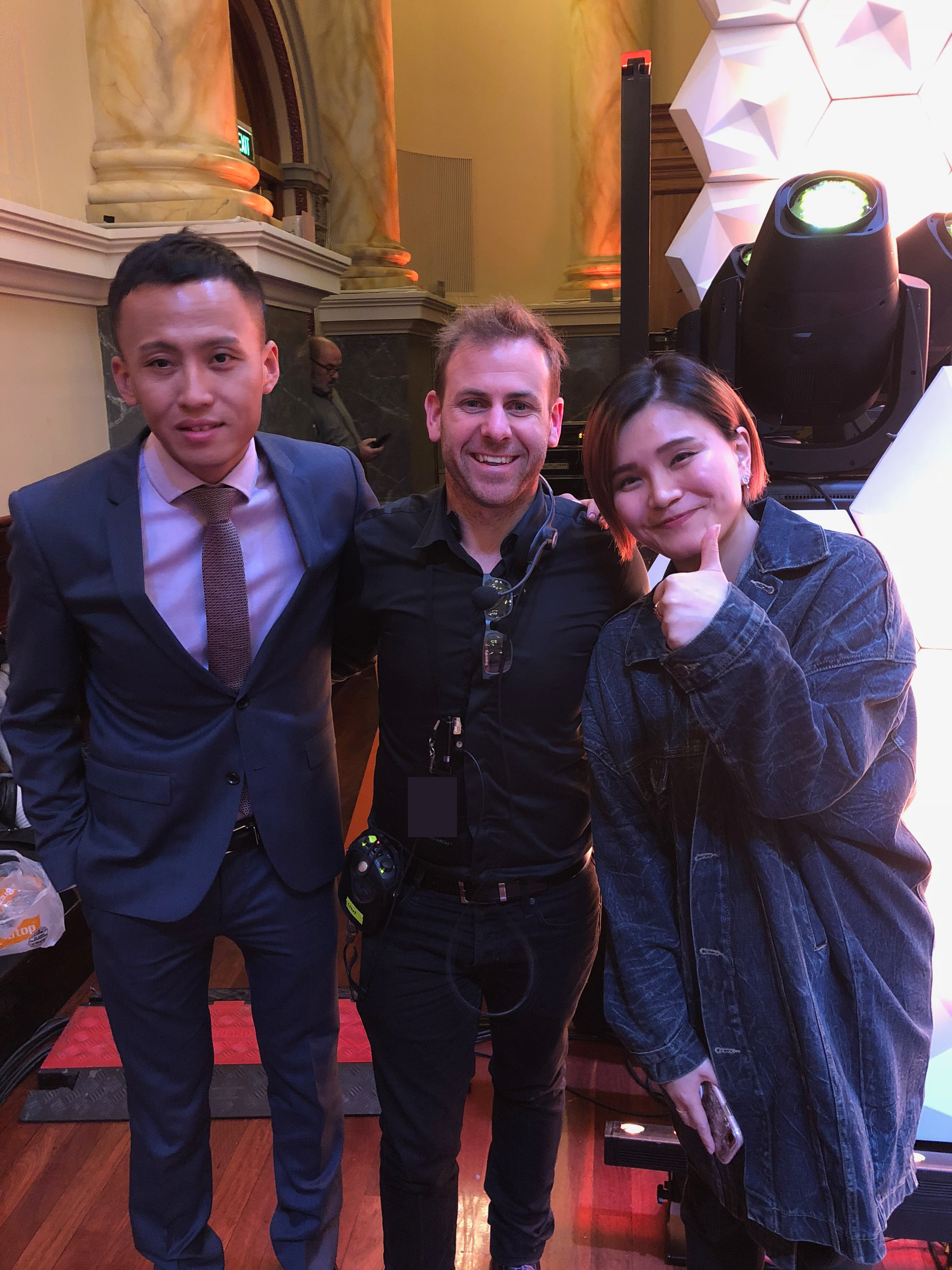 Three smiling people posing together in a room with ornate columns and modern lighting; two men and a woman, with one man wearing a suit and tie, the other man with a headset and equipment, and the woman giving a thumbs-up.