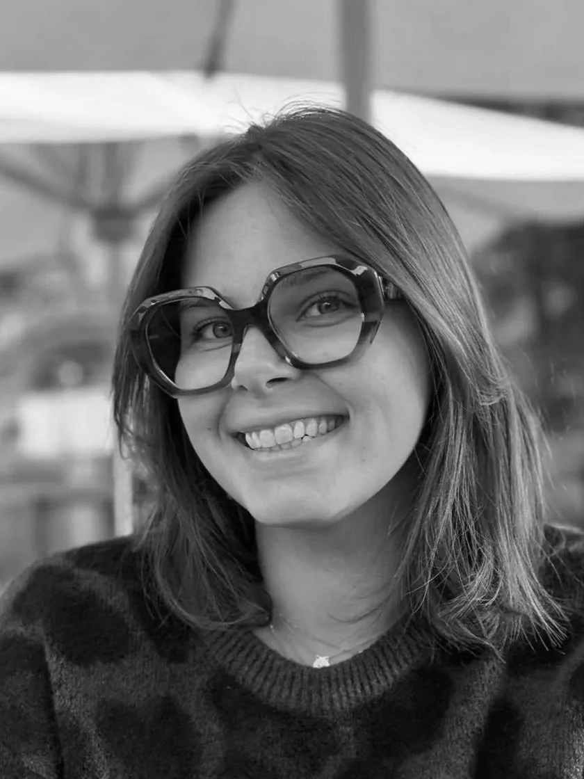 Une femme souriante porte des lunettes épaisses dans un environnement extérieur.