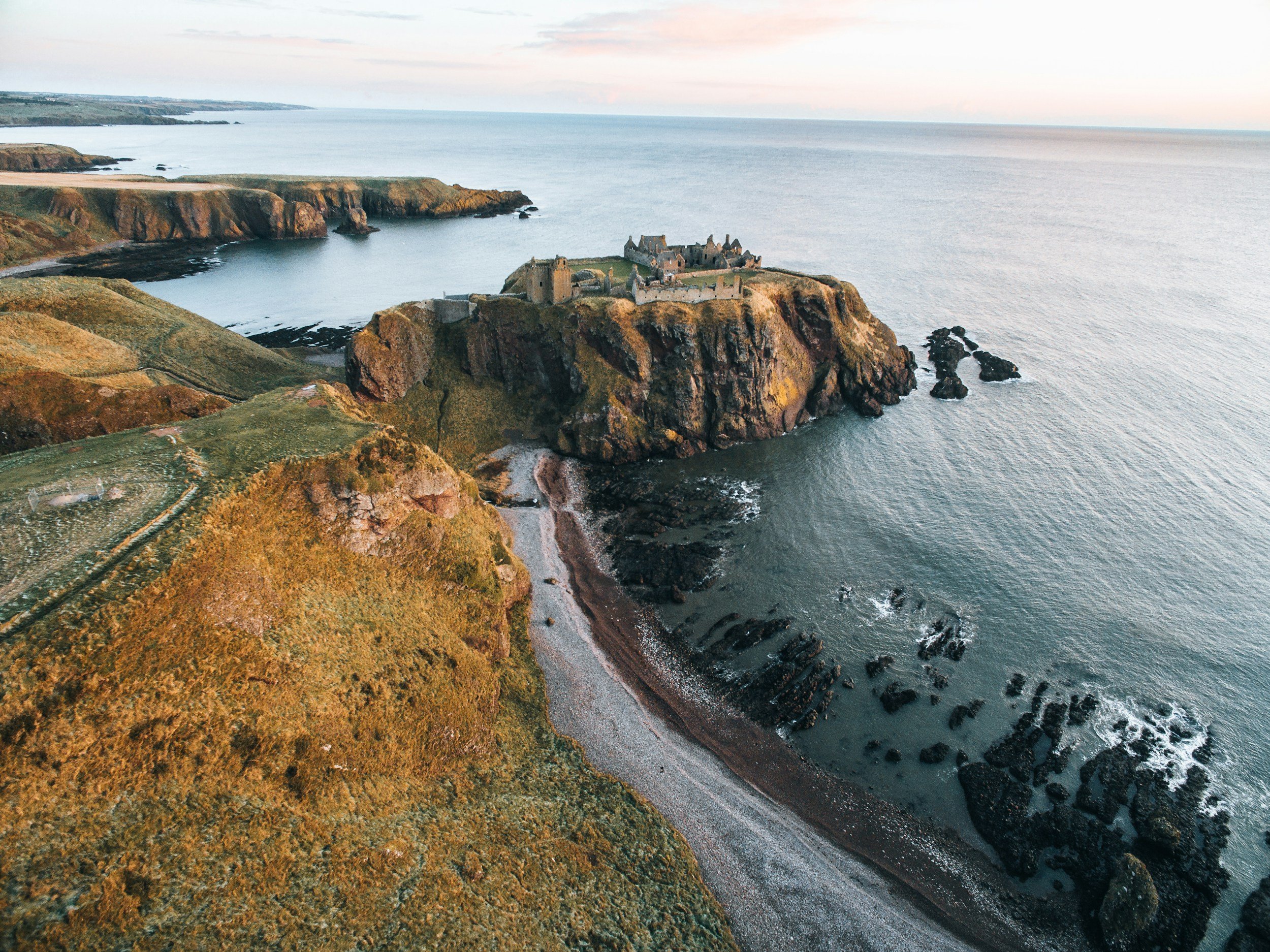 Aerial view of Dunluce Castle situated on a rocky cliff overlooking the ocean, with rugged cliffs and a sandy beach coastline.