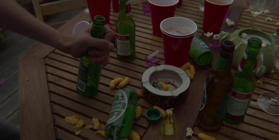 Empty beer bottles, red plastic cups, a green bottle, a brown bottle, an ashtray, and scattered popcorn on a wooden table at a party.