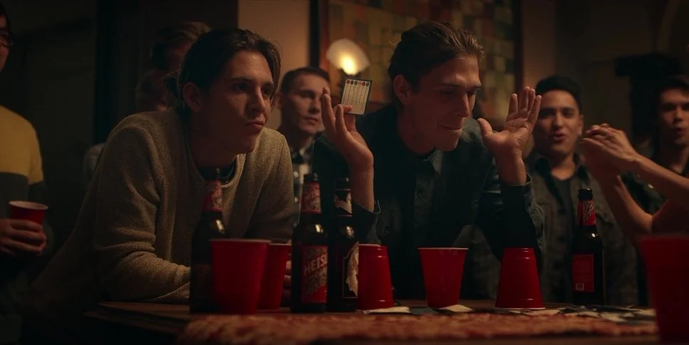 Young man with long hair sitting at a table with red cups and beer bottles, looking serious, in a dimly lit room with other people in the background. Another man with short hair is holding a card behind his head.