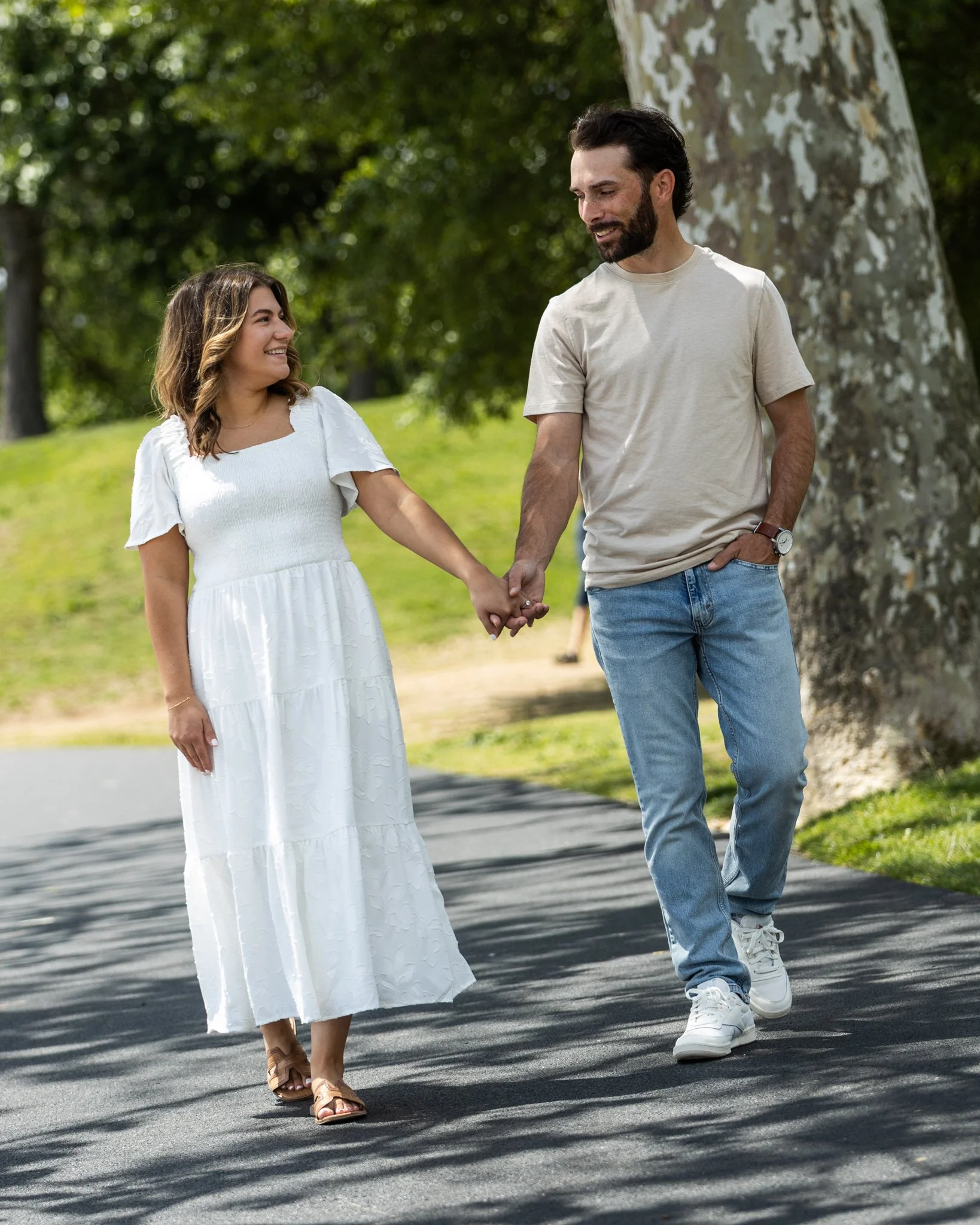 A couple walking hand in hand outdoors on a sunny day, smiling at each other, with trees and green grass in the background.
