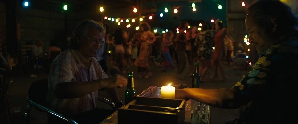 People dancing at a party with colorful string lights, a woman sitting at a table with a candle, and a man reaching for a drink.
