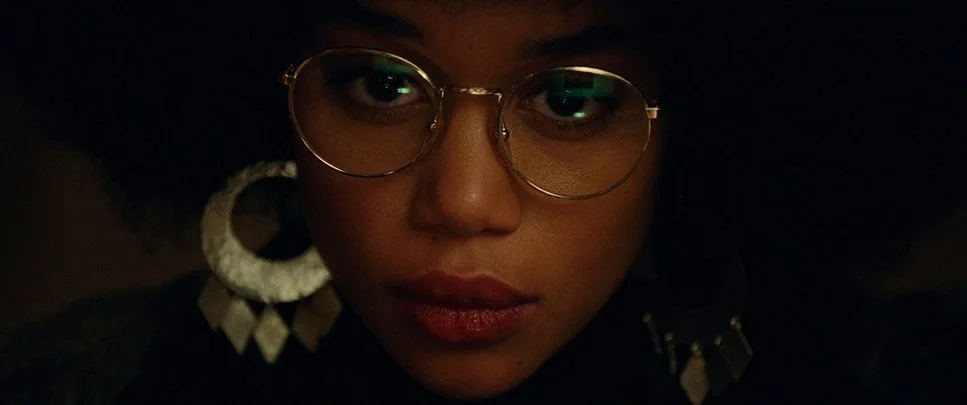 Close-up of a person's face wearing glasses and large earrings, with a dark background.