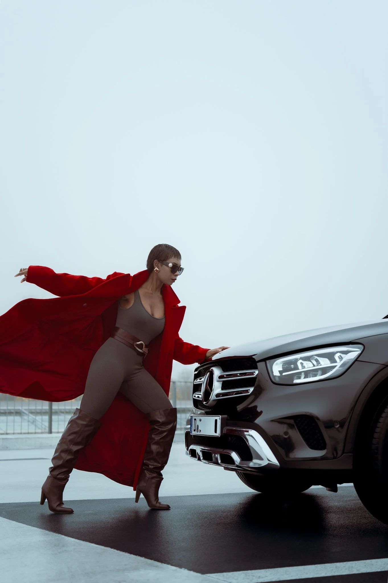 A woman dressed in a brown outfit and knee-high brown boots, wearing a long red coat and sunglasses, leaning on a black Mercedes-Benz car.