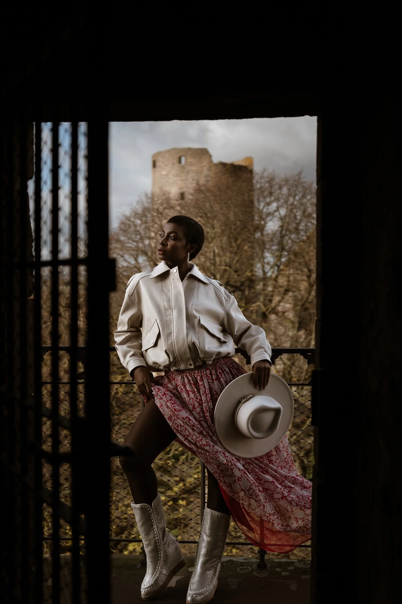 A woman standing on a balcony holding a hat, wearing a white jacket, patterned skirt, and silver boots, with an old castle and trees in the background.