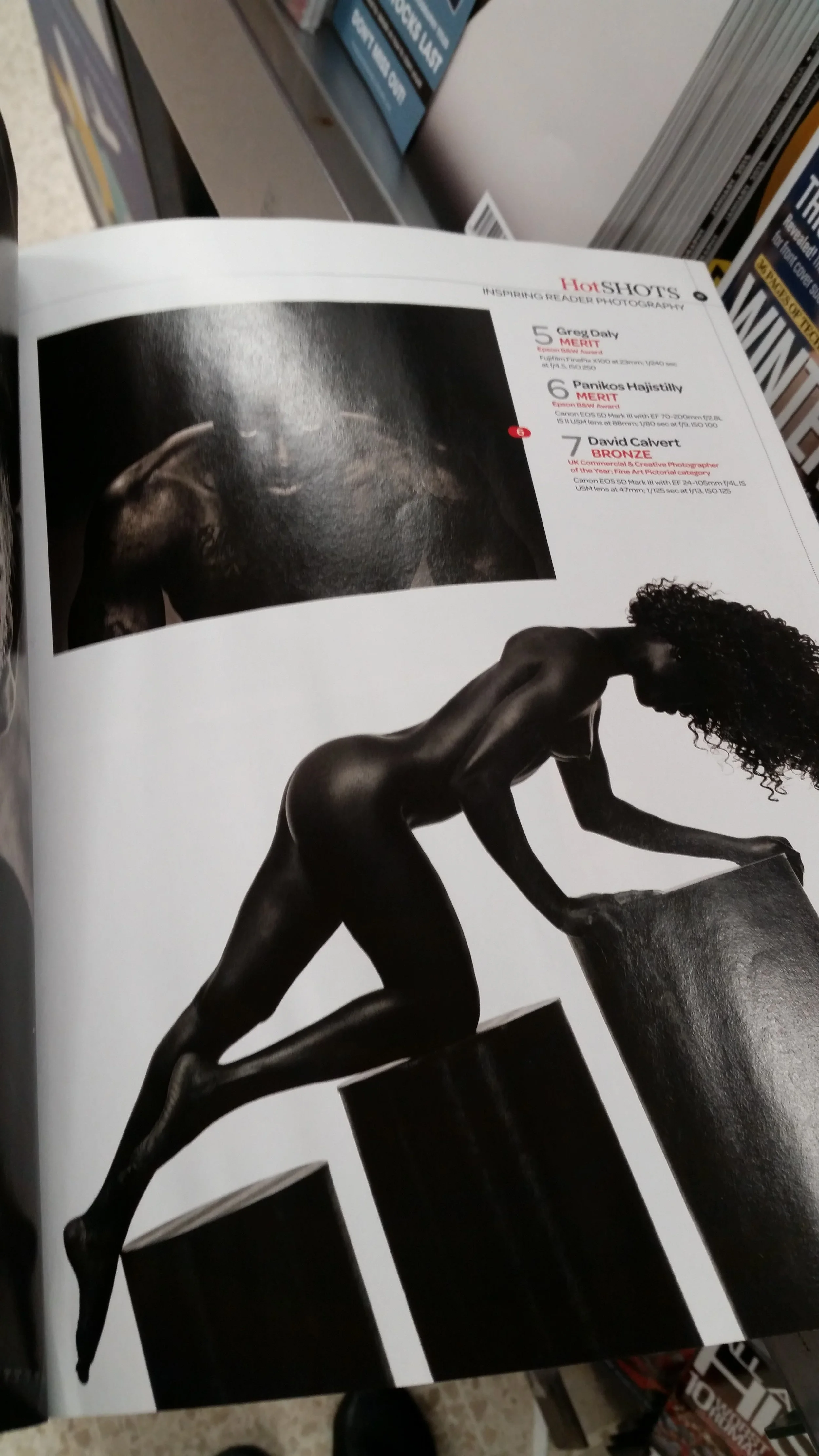 Black and white photograph of a nude woman with curly hair, leaning forward with her hands on large, rectangular black blocks. The woman appears to be posed as if climbing or pushing the blocks.