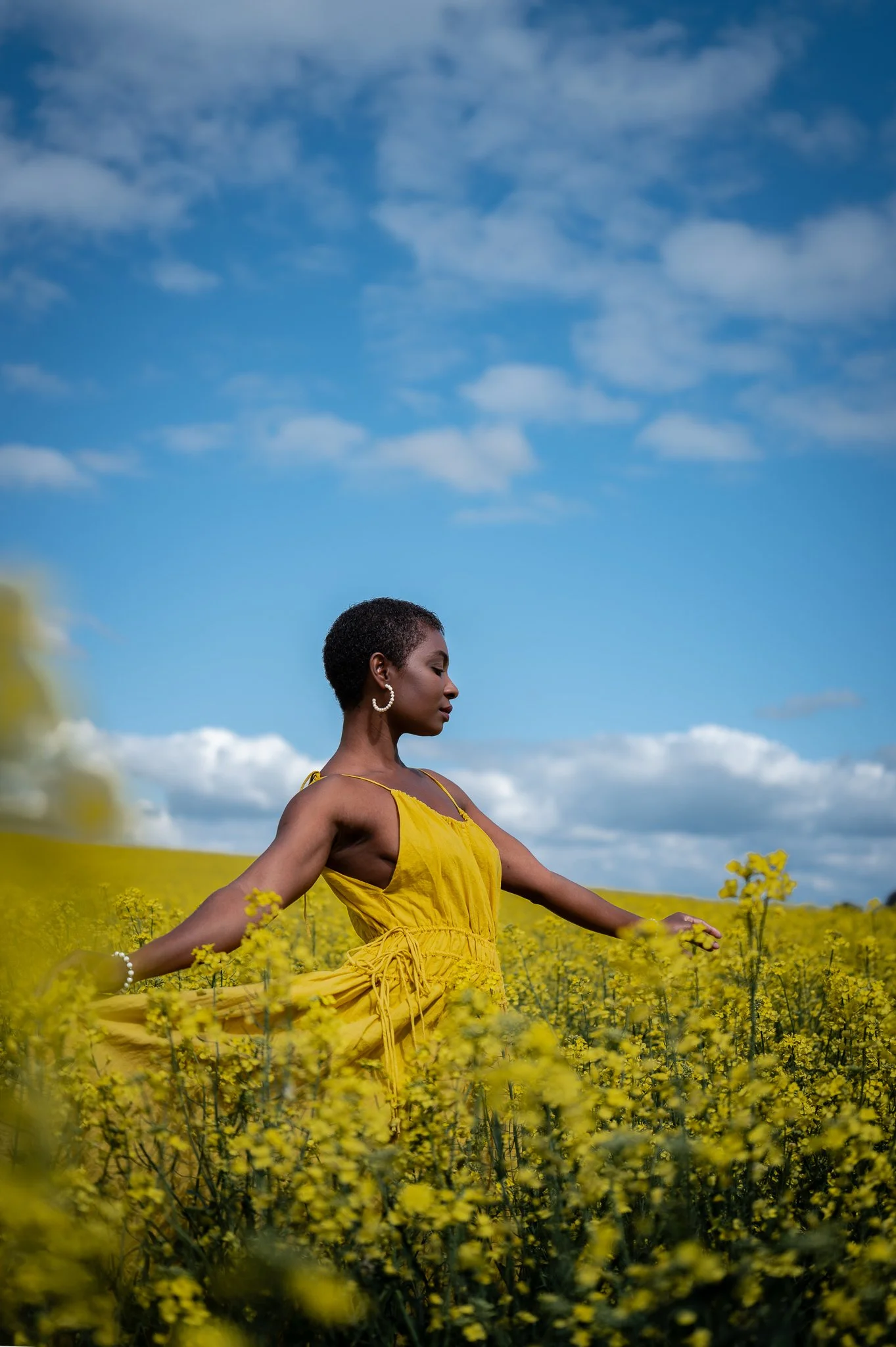 A woman in a yellow dress standing in a yellow flower field against a blue sky with clouds.