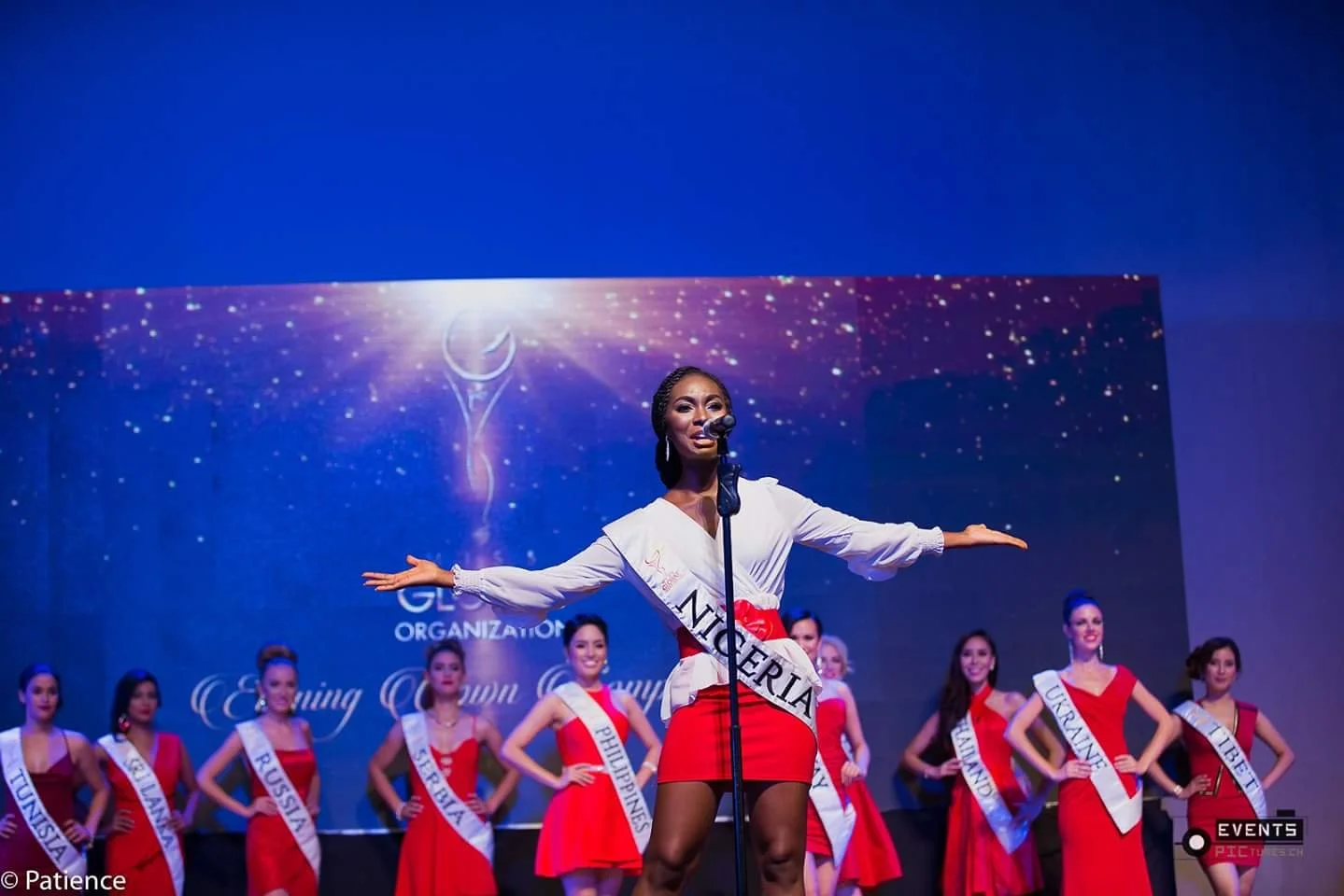 Beauty pageant contestants in evening gowns on stage, with one woman singing into a microphone in the foreground, wearing a sash that says 'NIGERIA'.