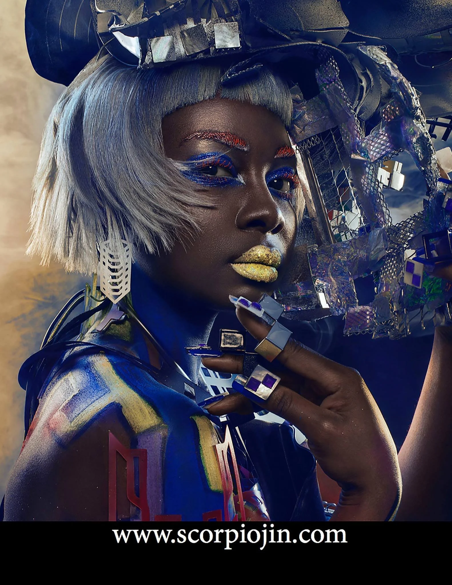 A person with futuristic, colorful makeup, silver wig, and elaborate accessories, posing with a creative, metallic, and holographic headpiece and clothing.