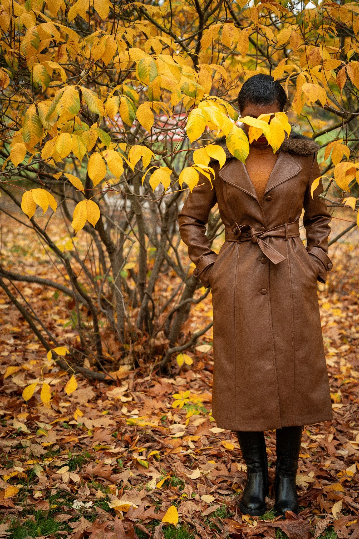 A woman in a long brown leather coat and black boots standing outdoors in autumn leaves, partially obscured by yellow leaves on a tree.