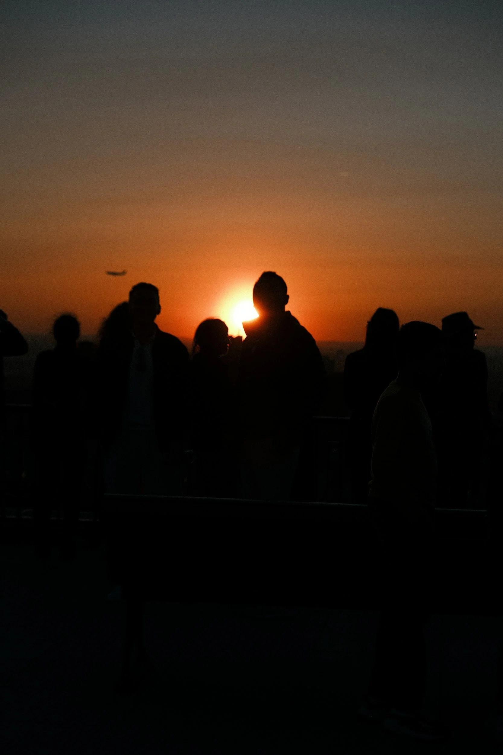 Silhouetted people standing on a beach at sunset, with an airplane flying in the sky.