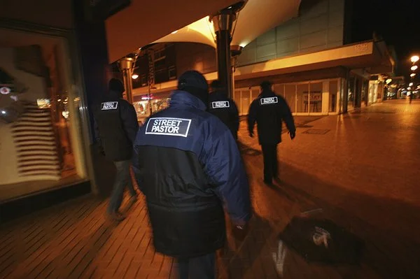 Four police officers walking outside a building at night, one with a jacket labeled "street pastor," with a suitcase on the ground.