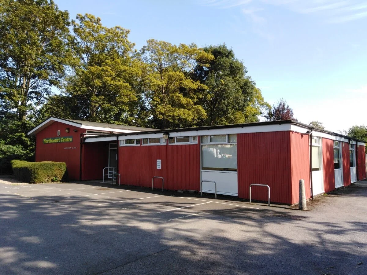 Red building with the sign 'Northcourt Centre' located near a parking lot with trees in the background.