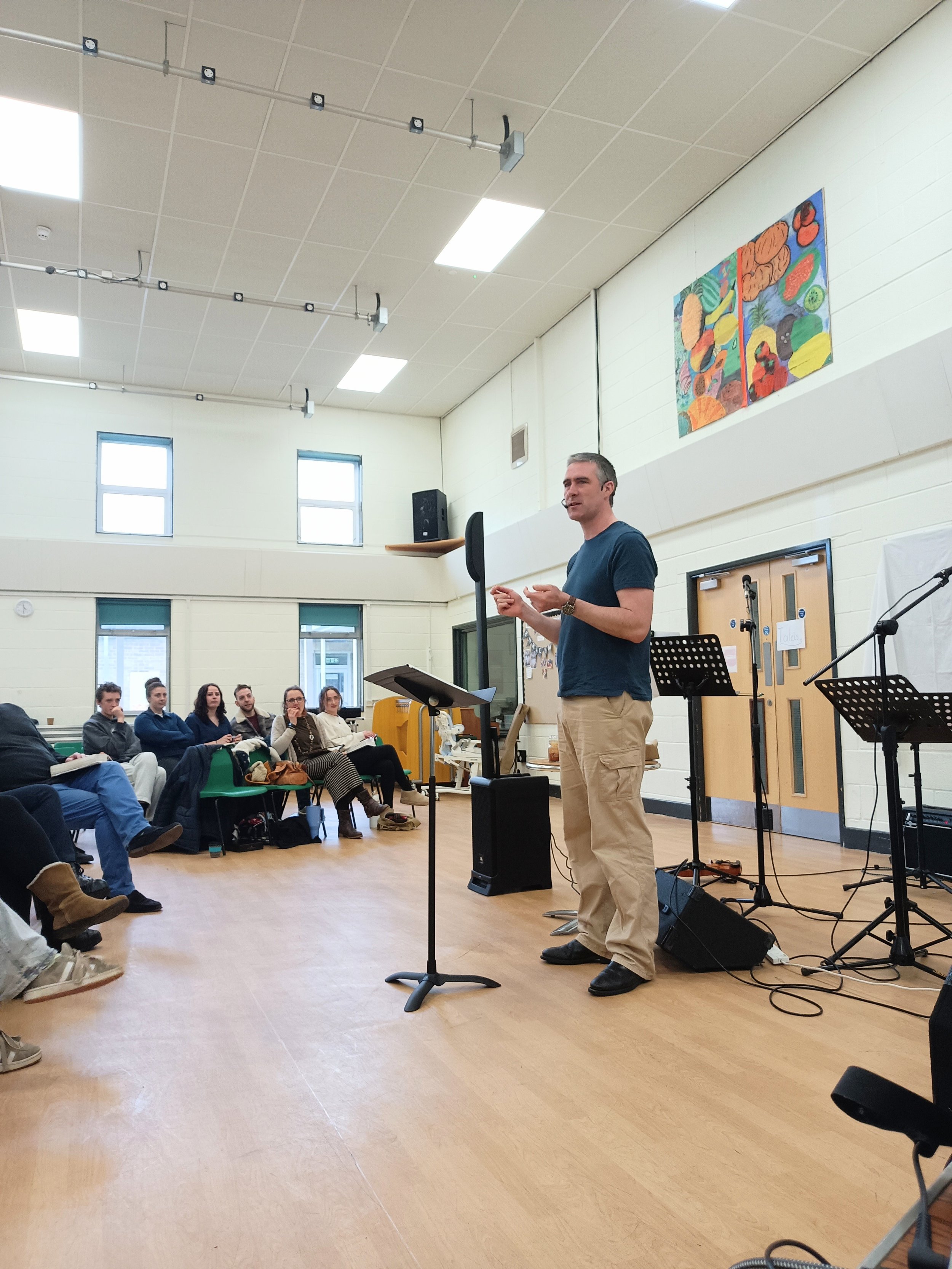 A man giving a presentation to an audience in a well-lit room with light-colored walls and wooden floors. There are musical stands, microphones, and a music stand near the man. An abstract painting is on the wall behind him, and several windows are on the upper part of the wall.