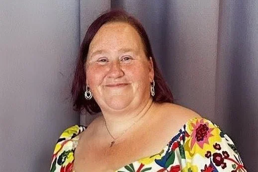 A woman with shoulder-length brown hair, wearing a colorful floral dress and earrings, smiling in front of a purple background.