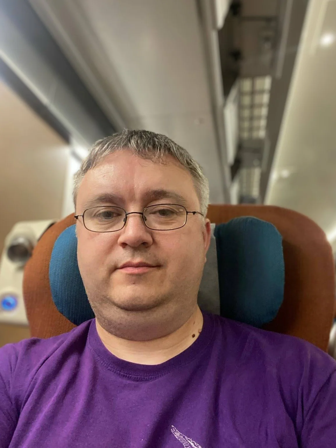 A man with glasses and short gray hair, wearing a purple shirt, sitting on a train with a headrest pillow behind him.