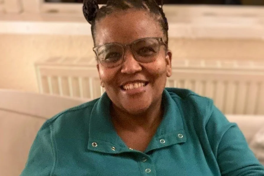 A woman smiling, wearing glasses and a teal collared shirt, sitting indoors with a radiator in the background.