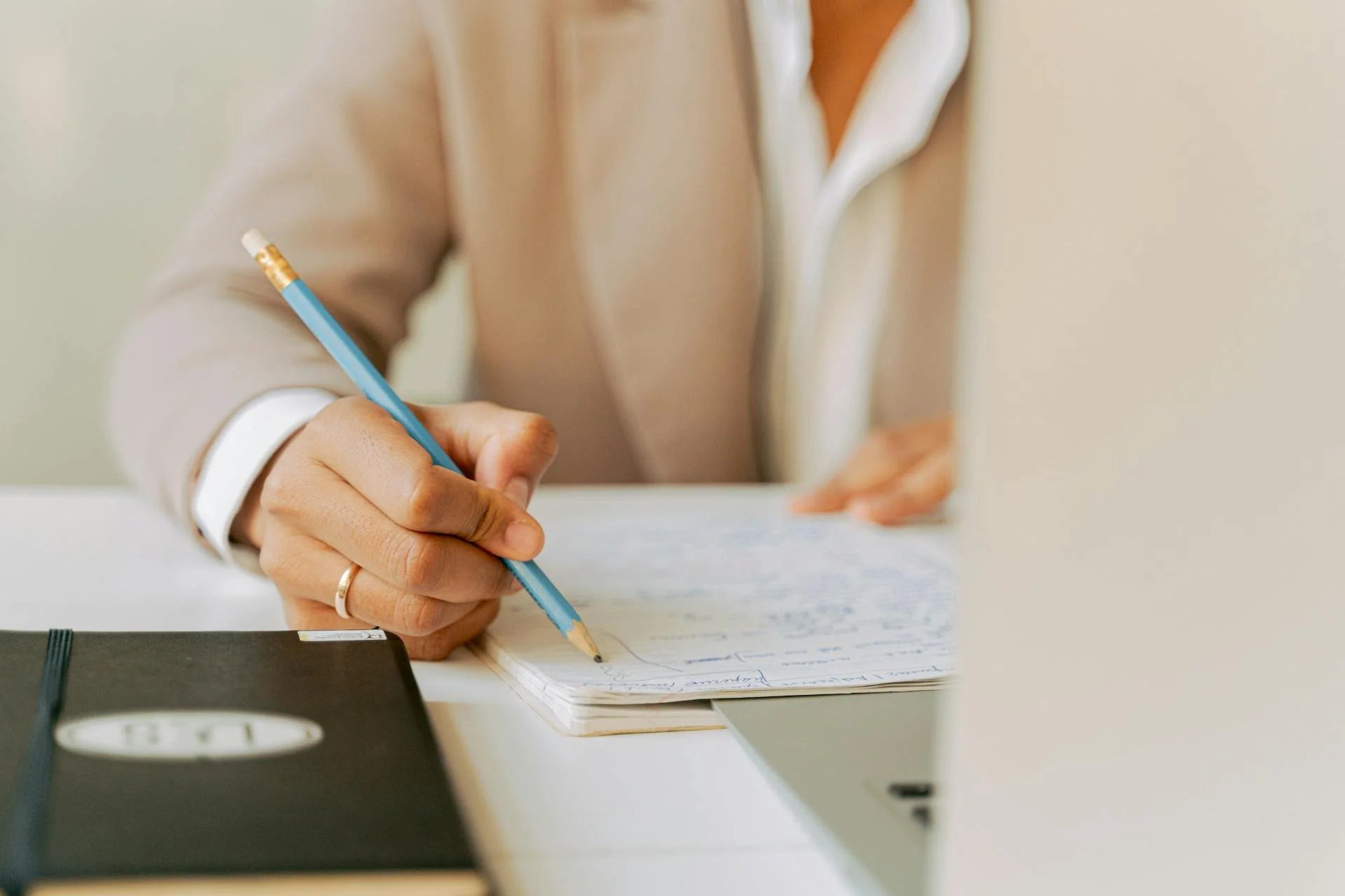 Een persoon schrijft in een schrift met een blauwe balpen, op een bureau met een zwarte agenda en een zwart apparaat, mogelijk een blokkeer of een oplaadpad.