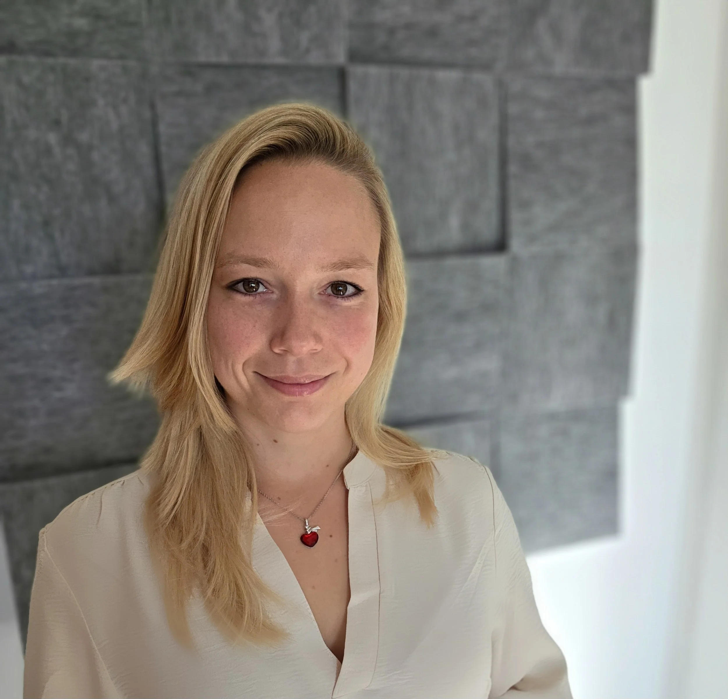 Porträt einer lachenden jungen Frau mit blonden Haaren, die eine weiße Bluse trägt, vor einer grauen Steinwand.