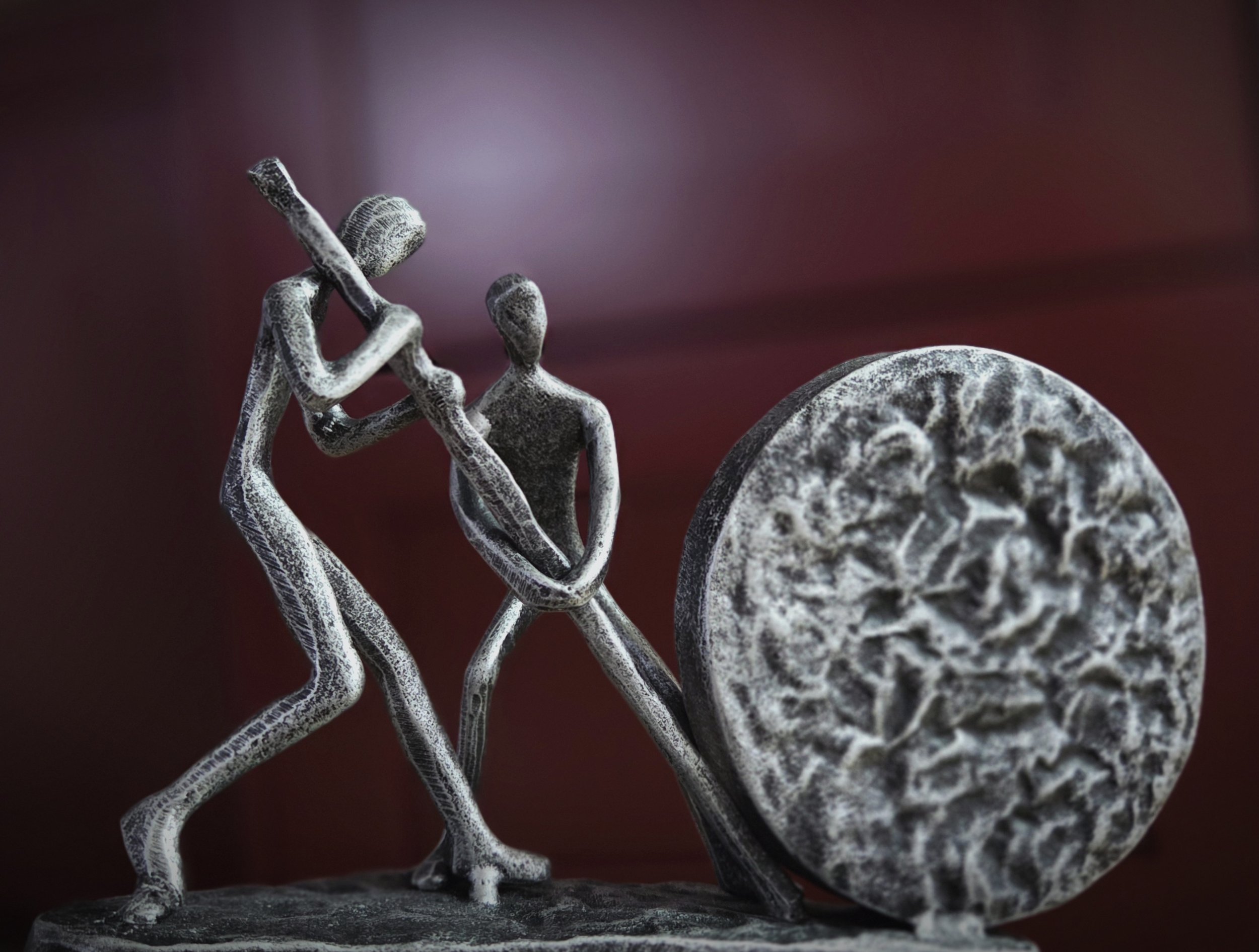 Skulptur von zwei stilisierten Menschen, die ein großes Rädchen schieben, vor einem dunkelroten Hintergrund.