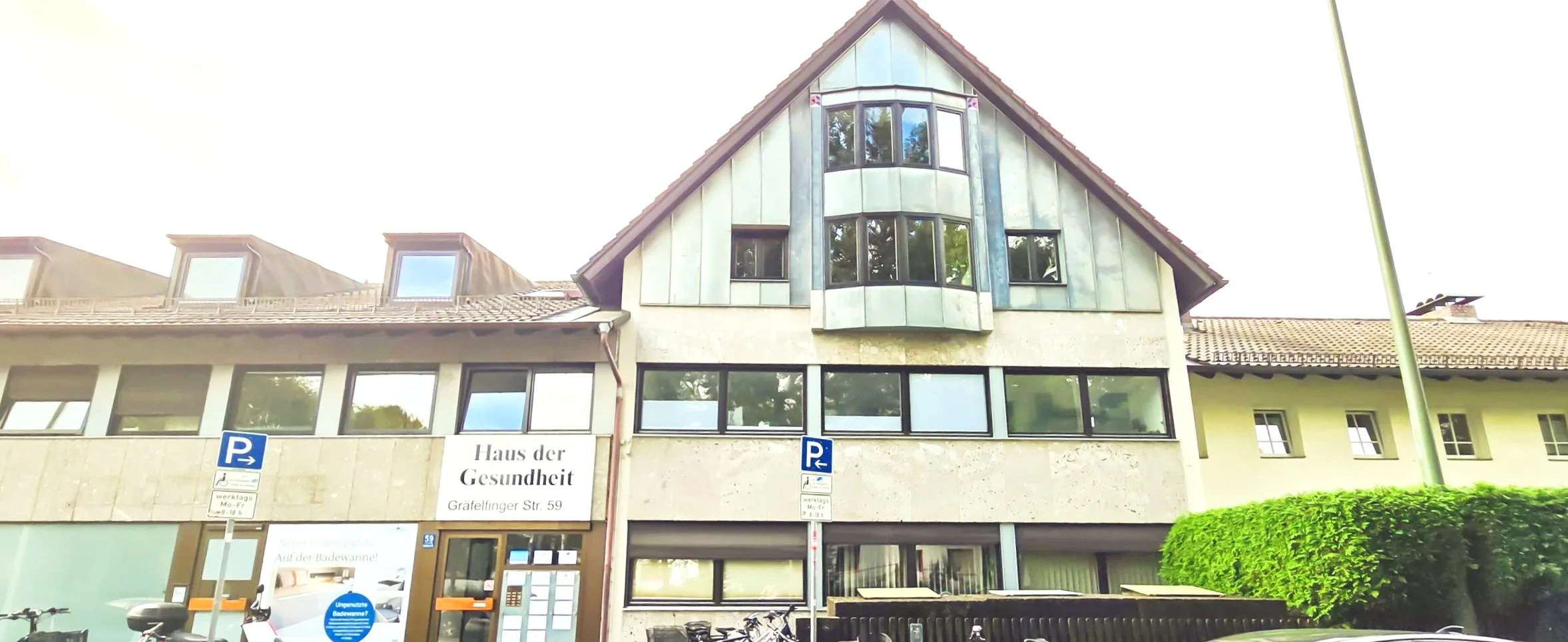 Mehrstöckiges Gebäude mit einem Dach und mehreren Fenstern, darunter ein Schild mit der Aufschrift 'Haus der Gesundheit' und Parkplätze davor.