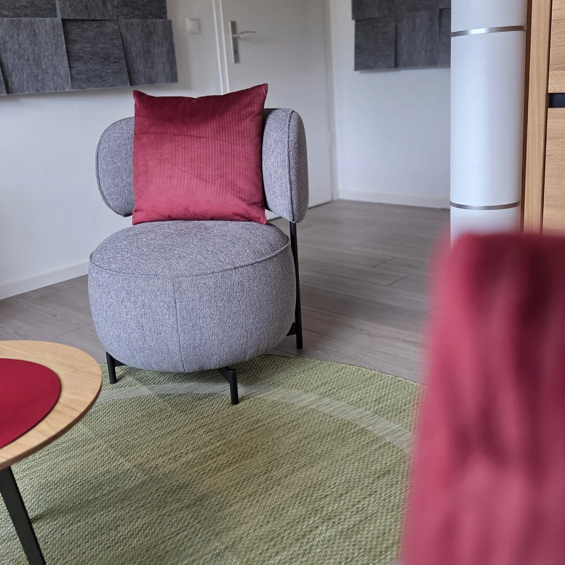 Ein modernes Zuhause mit einem grauen Sessel, rotem Kissen, grünem Teppich, Holztisch, weißen Wänden und einer Tür im Hintergrund.