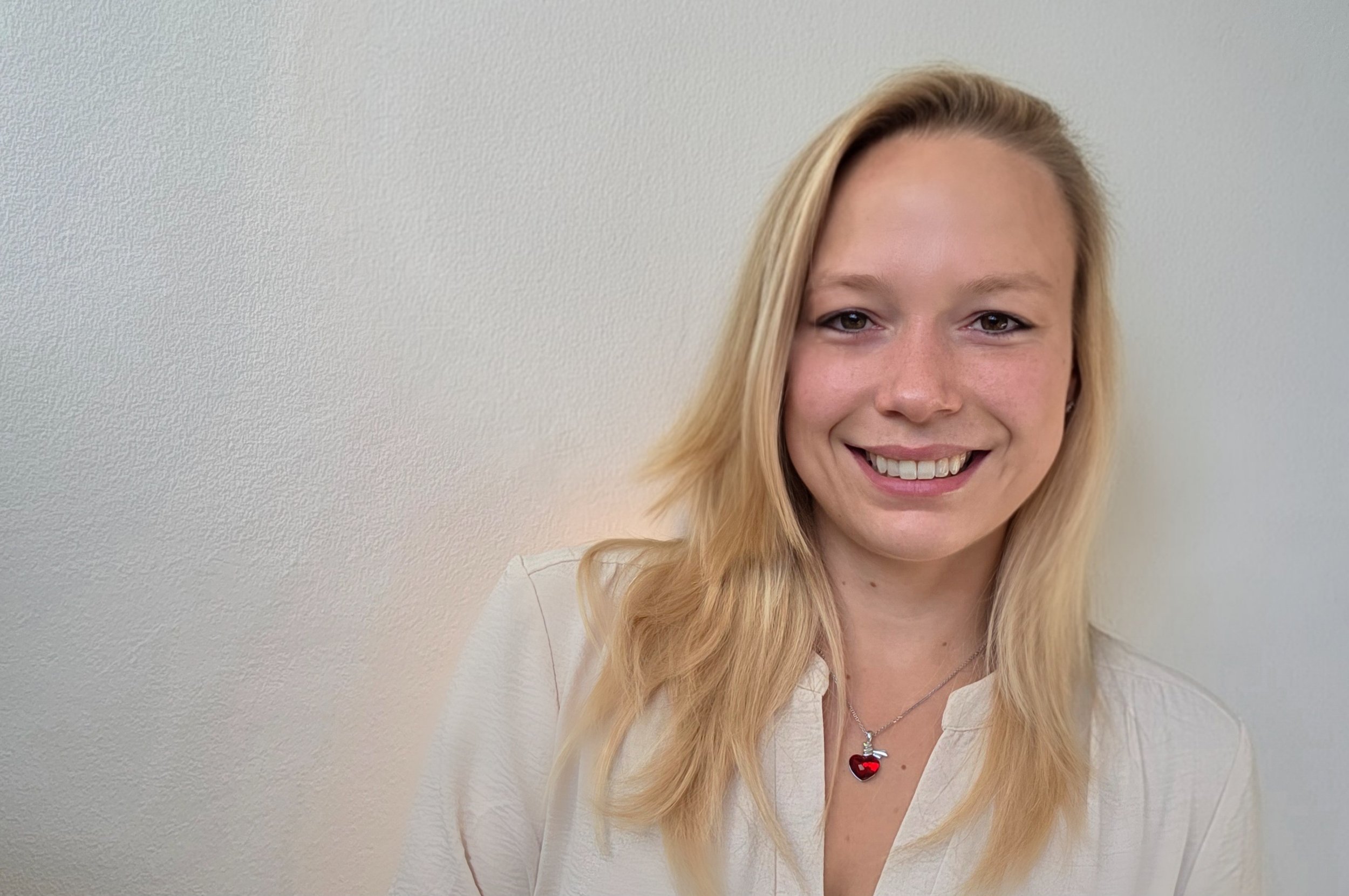 Ein Lächelndes junges Mädchen mit blonden Haaren, das eine weiße Bluse und eine Kette mit einem Herz-Anhänger trägt, vor einer hellen Wand.
